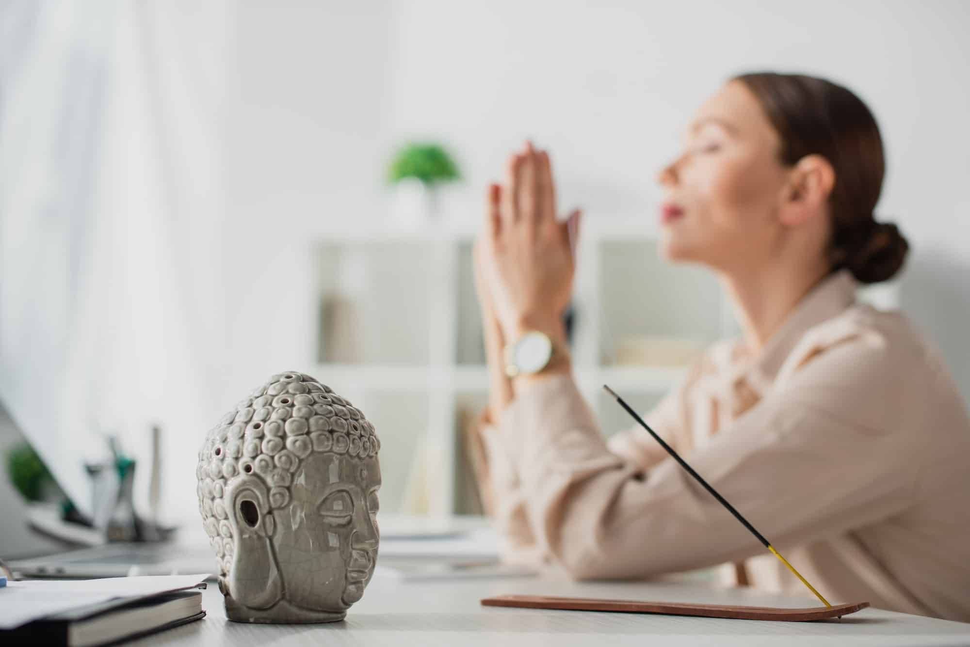 selektiver Fokus einer meditierenden Geschäftsfrau mit Namaste-Geste am Arbeitsplatz mit Buddha-Kopf und
